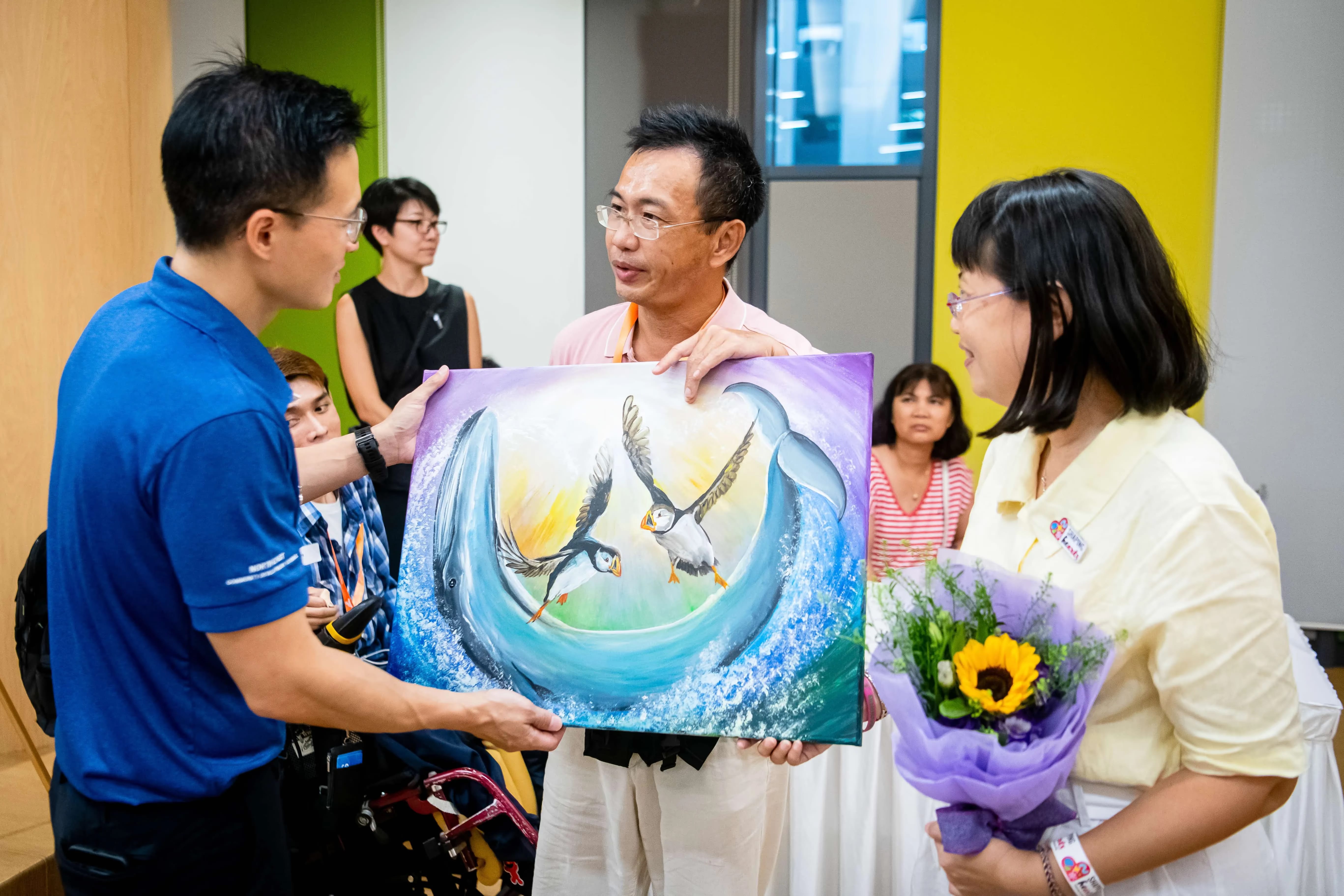 People hold a painting depicting dolphins and puffins as one woman stands holding a bouquet of flowers.